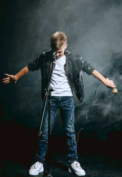 portrait of caucasian teenager in white t-shirt, blue jeans and leather jacket with microphone singing on dark background. hobby and glory concept - singing singer teenager contest imagens e fotografias de stock