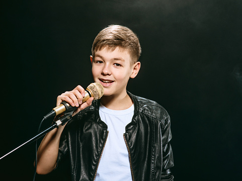 Portrait of caucasian teenager in white t-shirt, blue jeans and leather jacket with microphone singing on dark background. Hobby and glory concept