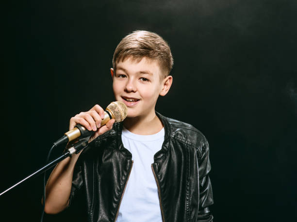 portrait of caucasian teenager in white t-shirt, blue jeans and leather jacket with microphone singing on dark background. hobby and glory concept - singing singer teenager contest imagens e fotografias de stock
