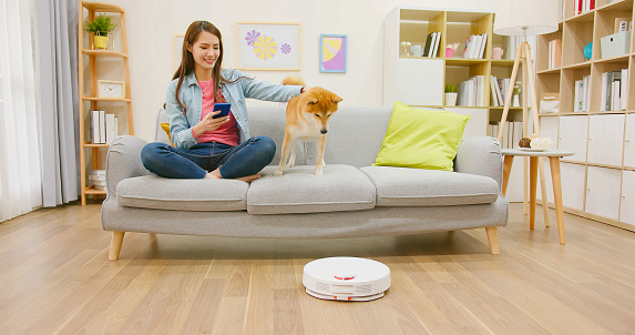 smart home concept - Happy beautiful young asian woman using robot cleaner sweeps floor with her dog at home