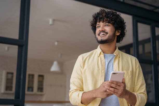 Smiling Indian man holding mobile phone shopping online looking away at home, copy space stock photo