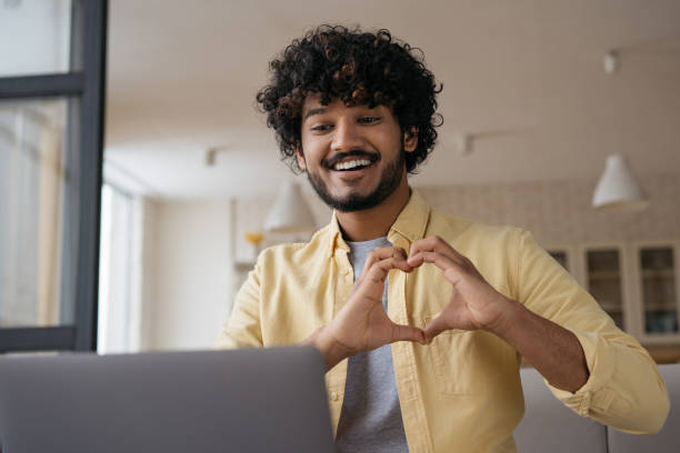 Indian influencer recording video communication online with subscribers showing heart symbol with love. Happy asian man using laptop computer having video conference sitting at home stock photo