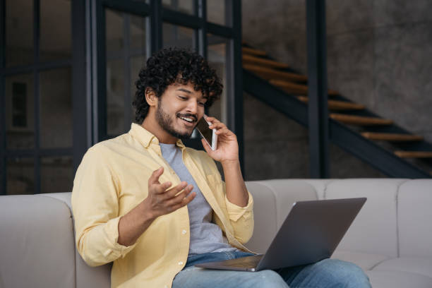 istock Indian business man talking on mobile phone planning start up sitting at workplace. Smiling freelancer using laptop holding smartphone, communication, working from home. Successful business 1384481966