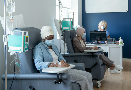 Two senior women sit side-by-side in their own chairs as they receive their cancer treatments intravenously. They are both dressed casually and have head scarves on as they sit looking across the room. A nurse can be seen working at a desk in the background.