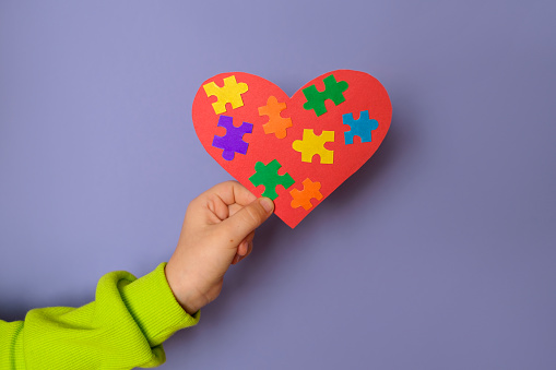 A big red heart with details of colored puzzles inside in a child's hand there is a symbol of autism. World Autism Day