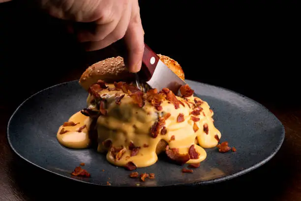 Corranso with knife a burger with melted cheddar cheese and bacon slices on rustic dish on wooden background