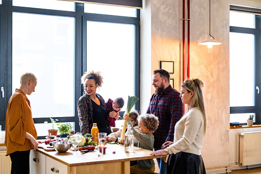Group of friends with their children talking while preparing food at kitchen island. Two couples with their kids sitting at kitchen table while cooking dinner at home.