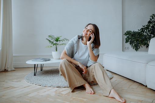 Smiling young brunette girl in a white t-shirt and beige pants sits on the floor in the living room at home and talks on a smartphone