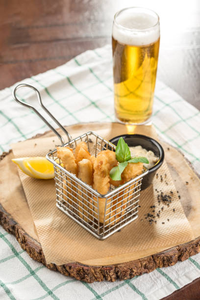 A basket of breaded fish sticks with sauce and glass of beer on wooden slab on the table stock photo