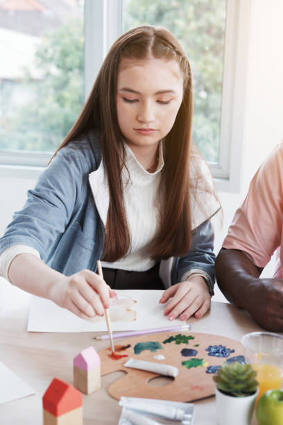 笑顔の白人のスタデンの女の子は、インターナショナルスクールの教室で絵筆と水彩画で絵を描いたり描いたりしています。アーティストコンセプトのための教育。 - group of people people family multi ethnic group ストックフォトと画像