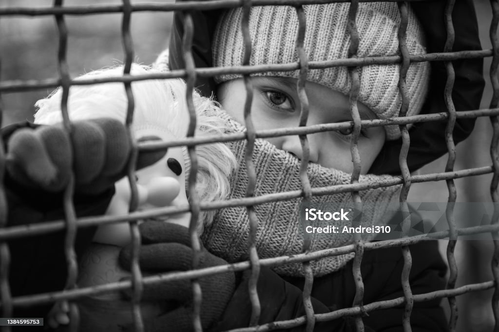 Little refugee girl with a toy behind a metal fence. Social problem of refugees and internally displaced persons. Refugee Stock Photo