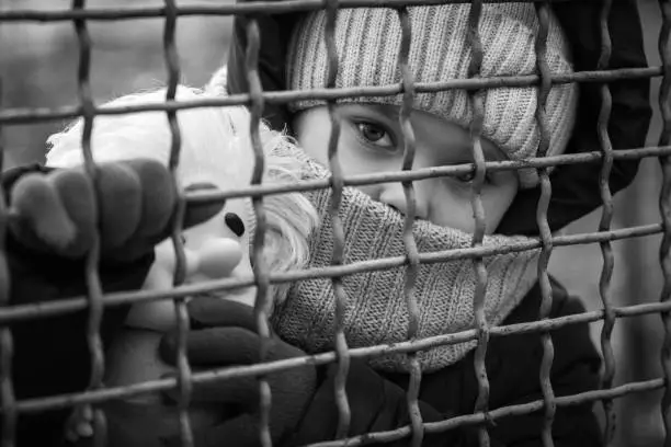 Photo of Little refugee girl with a toy behind a metal fence. Social problem of refugees and internally displaced persons.