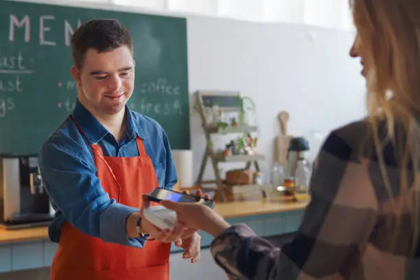 A cheerful young Down Syndrome waiter taking contactless smartphone payment from costumer in take away restaurant.