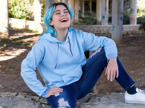 Laughing Blue haired Teenage girl in light blue hoodie sitting under bridge. Blue haired teen girl outdoors against bridge pillars. Clothing mockup