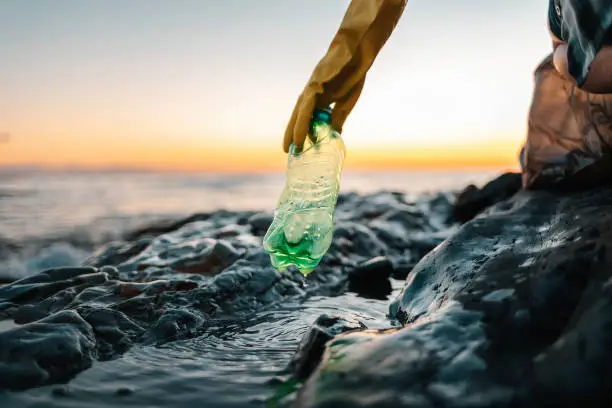 Environmental pollution. Volunteer in protective gloves picks up a plastic bottle on the beach. Close up of hand. Low angle view. Copy space. The concept of cleaning the coastal zone.