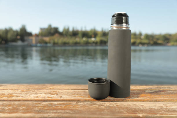 istock Modern grey thermos on wooden surface near river. Space for text 1384611987