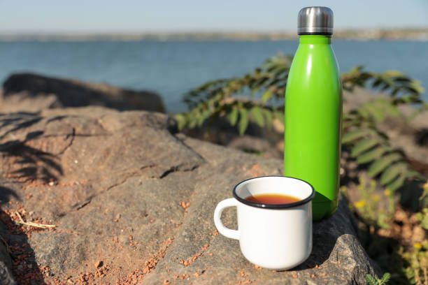istock Modern green thermos bottle and cup on stone near river. Space for text 1384613430