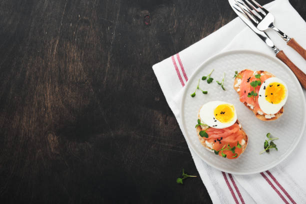 istock Sandwich with delicious toppings smoked salmon, eggs, herbs and microgreens radish, black sesame seeds over white plate on old wooden table background. Healthy open sandwich superfood. Top view. 1384613607