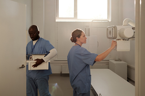 Pretty young clinician in medical scrubs preparing x-ray machine for examination of patients while her colleague leaving office