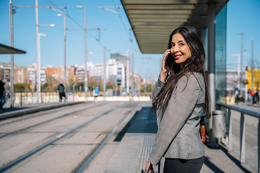 Side view of cheerful adult ethnic female entrepreneur with long dark hair in classy outfit smiling and looking at camera while talking on smartphone on tram stop in city
