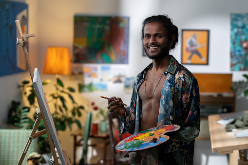 Happy young male artist with paintbrush and color palette looking at camera while standing in front of easel with unfinished painting