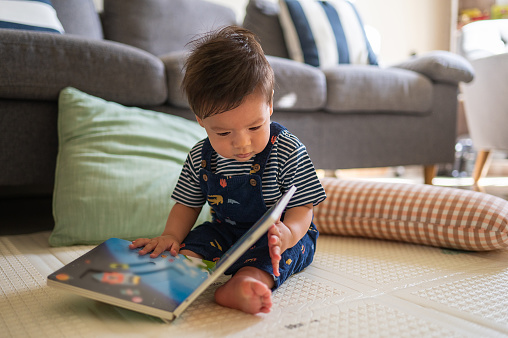 Infant mixed race baby boy playing like reading a book in the living room sitting on a baby safe soft playmat on the floor at home. Early education abstract