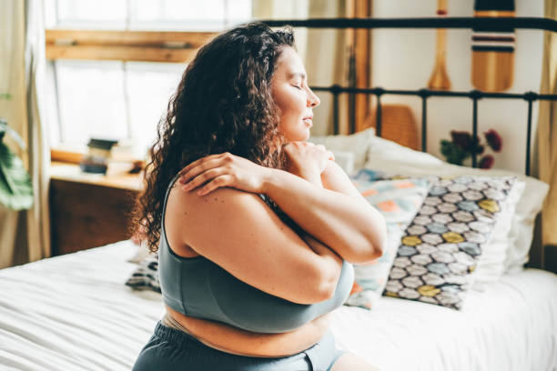 Curly haired overweight young woman in blue top and shorts with satisfaction on face accepts curvy body shape in stylish bedroom. stock photo