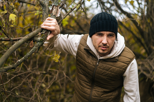 Close-up portrait of frozen survivalist male wearing warm clothes wading through thickets in forest in cold overcast day, looking at camera. Concept of exploration, travel and adventure in nature.
