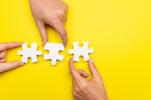 Three hands are holding three puzzle pieces next to another  in front of yellow background.