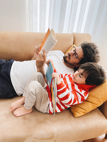 Father and son lying on sofa at home and reading book together