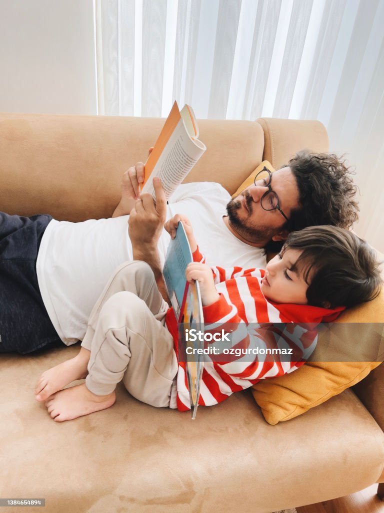 Father and son lying on sofa at home and reading book together Family Stock Photo