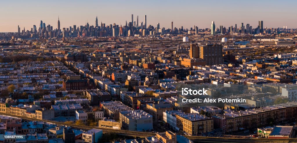 Manhattan Midtown Skyline includes the Empire State Building, Hudson Yards, and other iconic skyscrapers. View over the residential district of Bushwick, Brooklyn, at sunset.   Extra-large, high-resolution stitched panorama. Remote view of the Manhattan Skyline illuminated in the night over the residential district of Bushwick, Brooklyn, in the sunset. Brooklyn - New York Stock Photo