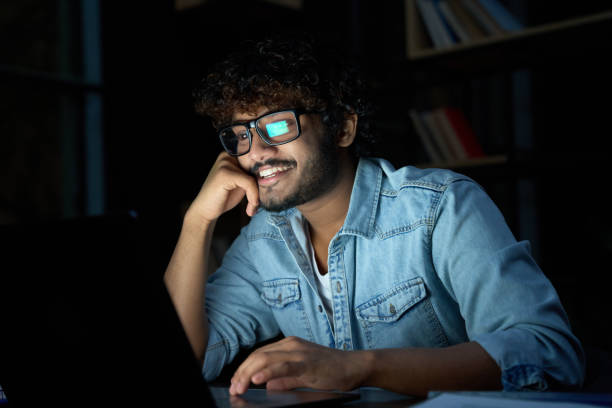 Happy young indian man working online late at night using laptop computer.