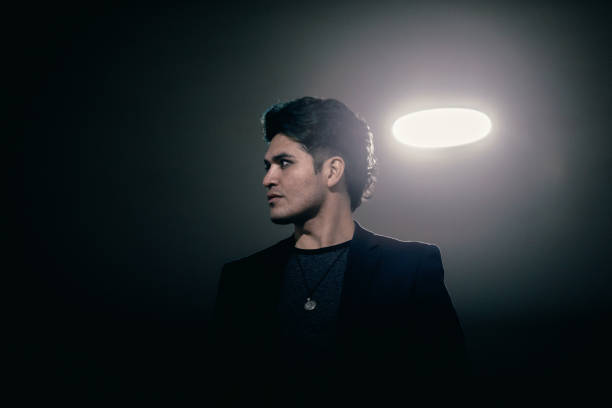 A Hispanic man in dress casual clothing stands in a dimly lit city parking lot, the overhead lights making for a high contrast portrait.