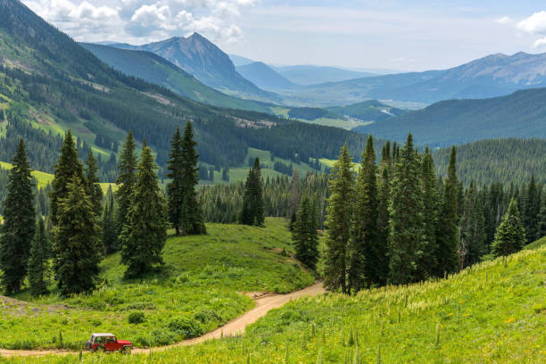 Mount Crested Butte - A red SUV running down a winding mountain road in a green valley towards Mount Crested Butte on a sunny Summer morning. Crested Butte, Colorado, USA. stock photo