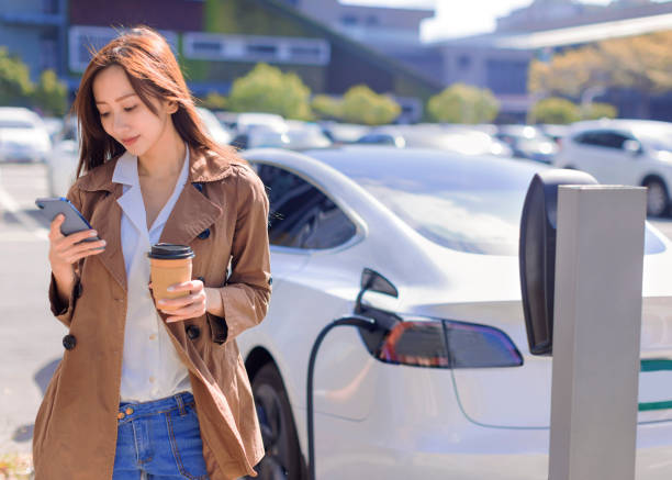 Smiling young woman standing on city parking near electric car, charging automobile battery from small city station, drinking coffee and using smartphone stock photo