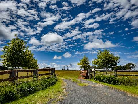 Horizontal landscape photo of the grass-lined gravel driveway and old wooden fences at the entrance to a rural farm in the New England high country near Armidale, NSW in Autumn. Atmospheric blue sky with white fluffy clouds.