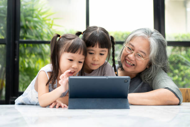 彼女の2人の孫とアジアの祖母は、リビングルームの自宅でデジタルタブレットでオンラインで教育ゲームを楽しんで遊んでいます。オンライン教育と親からの思いやりの概念。 - grandmother child senior adult multi generation family ストックフォトと画像