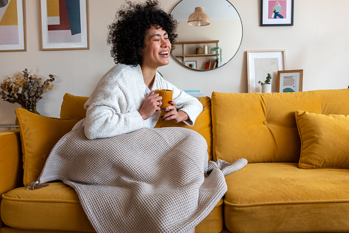 Happy African american woman enjoying quiet time at home laughing, drinking morning coffee sitting on sofa. Copy space