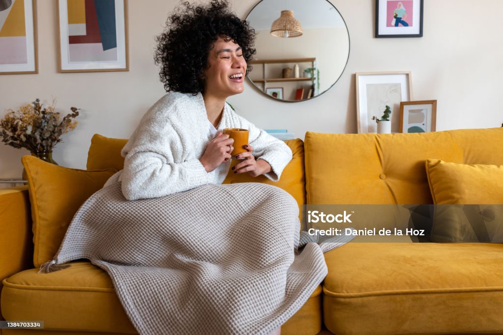 Happy African american woman enjoying quiet time at home laughing, drinking morning coffee sitting on sofa. Copy space Happy African american woman enjoying quiet time at home laughing and drinking morning coffee sitting on sofa. Copy space. Lifestyle concept. Winter Stock Photo