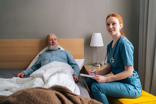 Positive female doctor writing diagnosis and symptoms in medical history, looking at camera, talking to senior male patient lying on bed at home. Elderly man getting medical consultation in hospital.