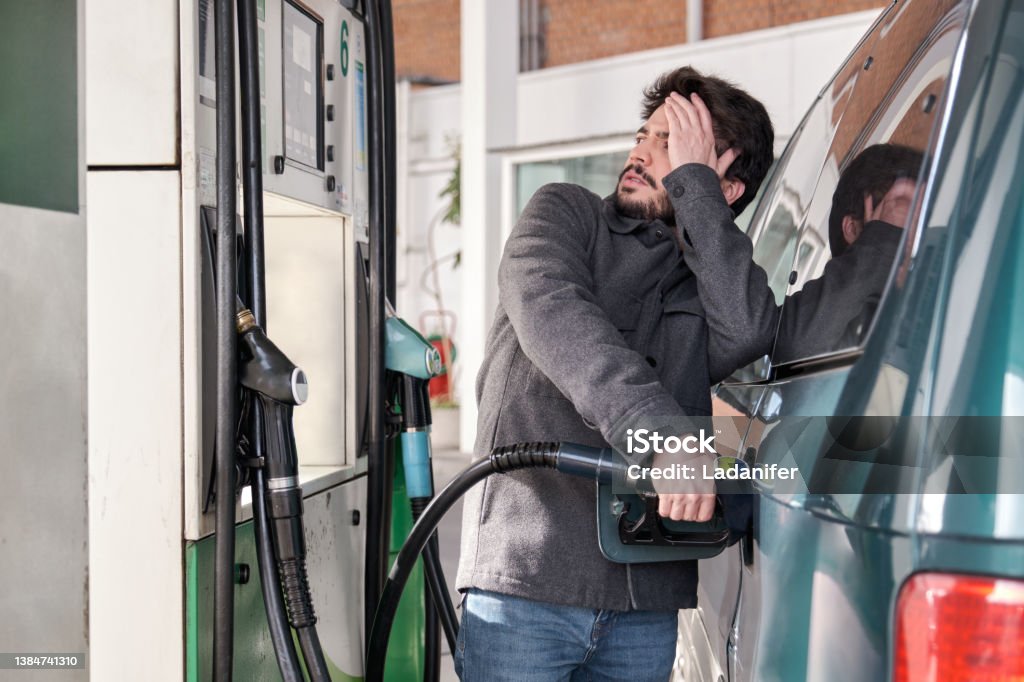 Young man refueling his vehicle while looking worried at the high gas prices. Young man refueling his vehicle while looking worried at the high gas prices at a gas station. Fuel Prices Stock Photo