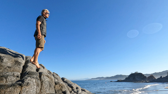 Happy ethnic guy in casual summer clothes smiling with backpack on shoulder while standing on stony sea coast in summer day