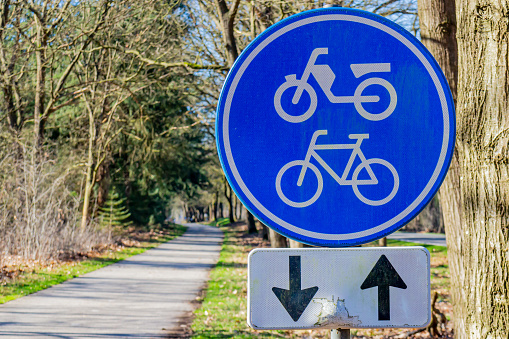 Traffic sign: Lane for bicycles and mopeds in both directions, blue and white round plate, cycle path between trees that is lost in the blurred background, sunny day in the Netherlands