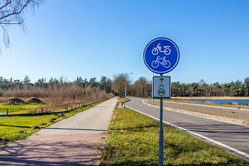 Bike lane next to a countryside road, sign indicating: bicycle and moped lane, route 98, lake with blue water and abundant trees in the blurred background, sunny day in North Brabant, Netherlands