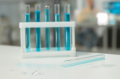 Shattered glass of broken test tube on desk against group of flasks with blue liquid in modern clinic or scientific laboratory
