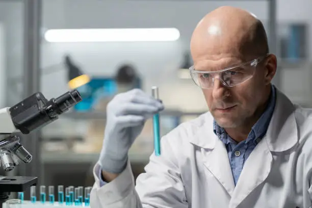 Mature serious researcher in gloves, eyeware and lab coat studying characteristics of blue liquid while holding flask in front of himself