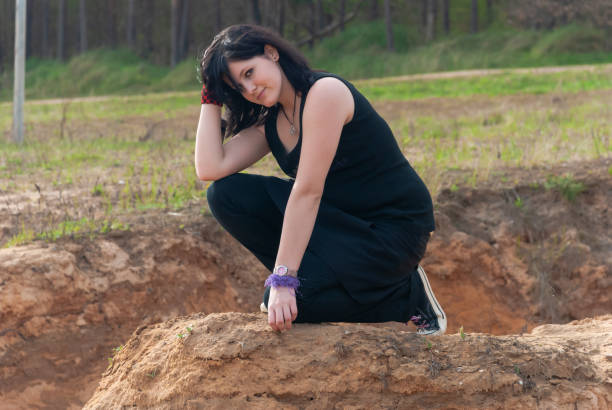 Punk emo girl, young adult with black hair, kneeling, looking at camera Punk emo girl, young adult with long black hair, kneeling on a hill outdoors in the nature, looking at camera, horizontal black hair emo girl stock pictures, royalty-free photos & images