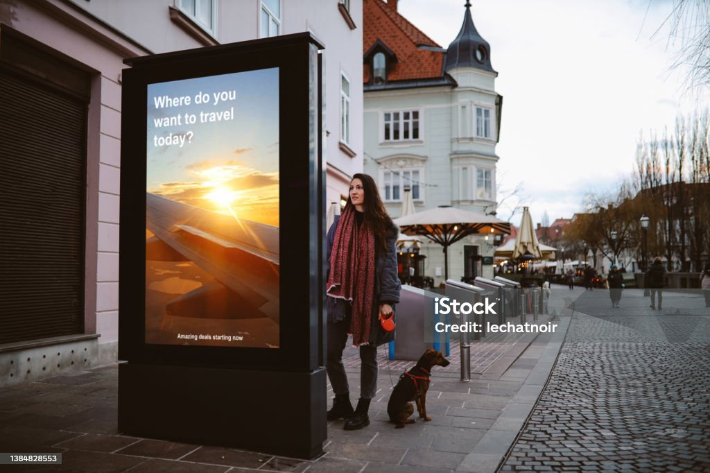 Is it time to travel again? Woman in her late 30s looking at the electronic smart sign in the Ljubljana city centre, browsing the tourist locations offers thinking of traveling again after the pandemic. Advertisement Stock Photo