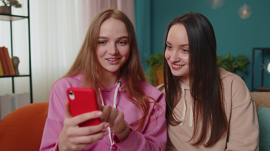 Cheerful girls friends siblings watching funny movies on smartphone, online comedy film. Happy two female women couple family on sofa at home, laughing, spend leisure time on social media network